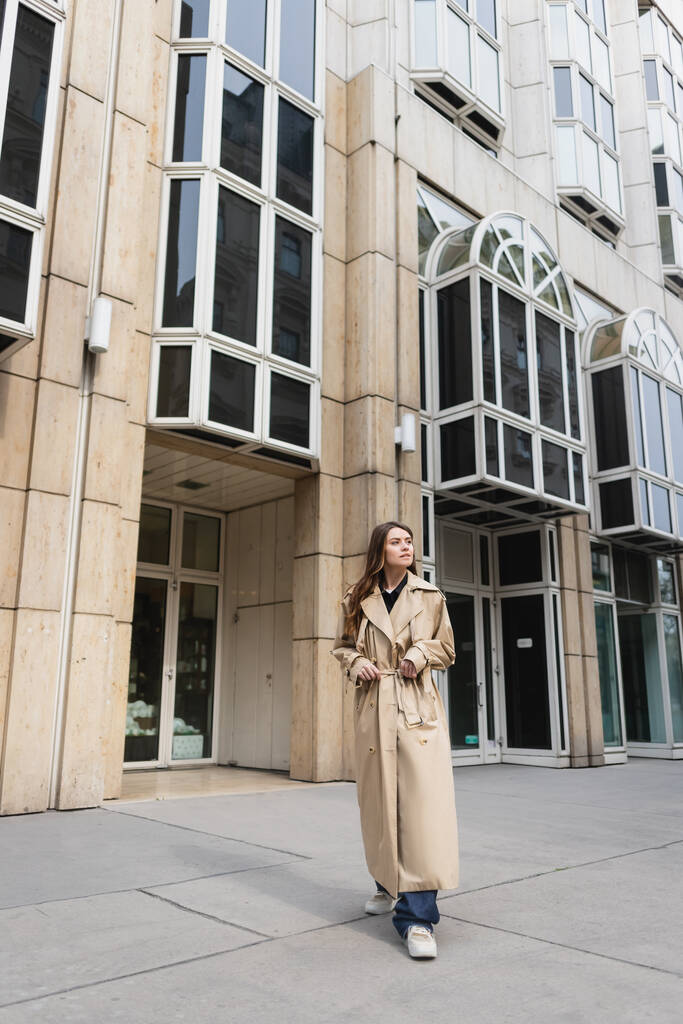 volledige lengte van jonge vrouw in stijlvolle trench jas staan in de buurt van gebouw in vienna  - Foto, afbeelding