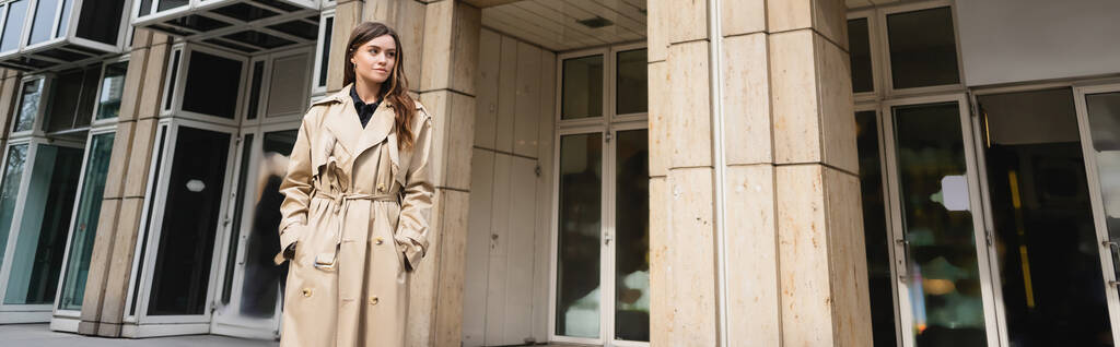 jovem mulher em casaco de trincheira elegante de pé com as mãos em bolsos perto do edifício em Viena, banner - Foto, Imagem