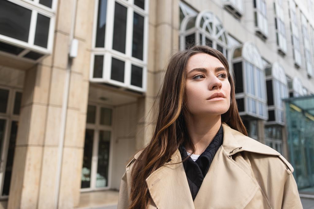 молода жінка в стильному тренч пальто дивиться далеко біля будівлі
 - Фото, зображення