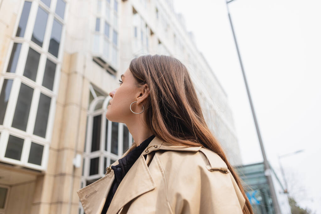 low angle view of young woman in beige trench coat looking at building - Photo, Image
