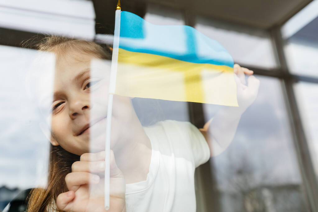 Mädchen mit der ukrainischen Flagge in den Händen schaut aus dem Fenster - Foto, Bild