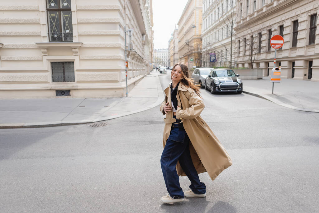 повна довжина веселої молодої жінки в бежевому траншеї пальто, що йде на європейській вулиці
  - Фото, зображення