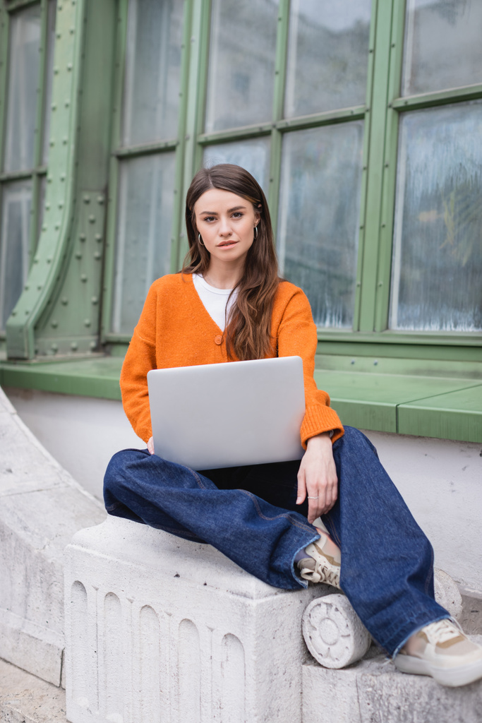 ジーンズとオレンジのカーディガンを着た若い女性がビルの屋上にノートパソコンを持って座り - 写真・画像