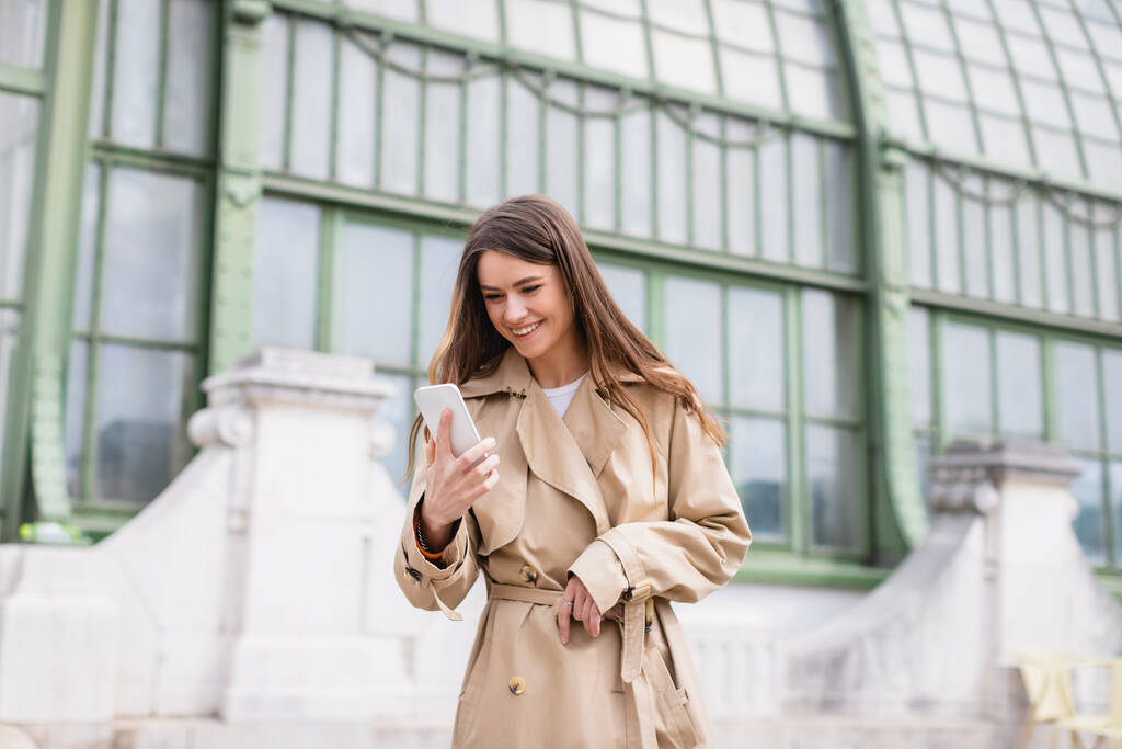 Ευτυχισμένη νεαρή γυναίκα με καμπαρντίνα κοιτάζοντας το smartphone κοντά στο ευρωπαϊκό κτίριο  - Φωτογραφία, εικόνα