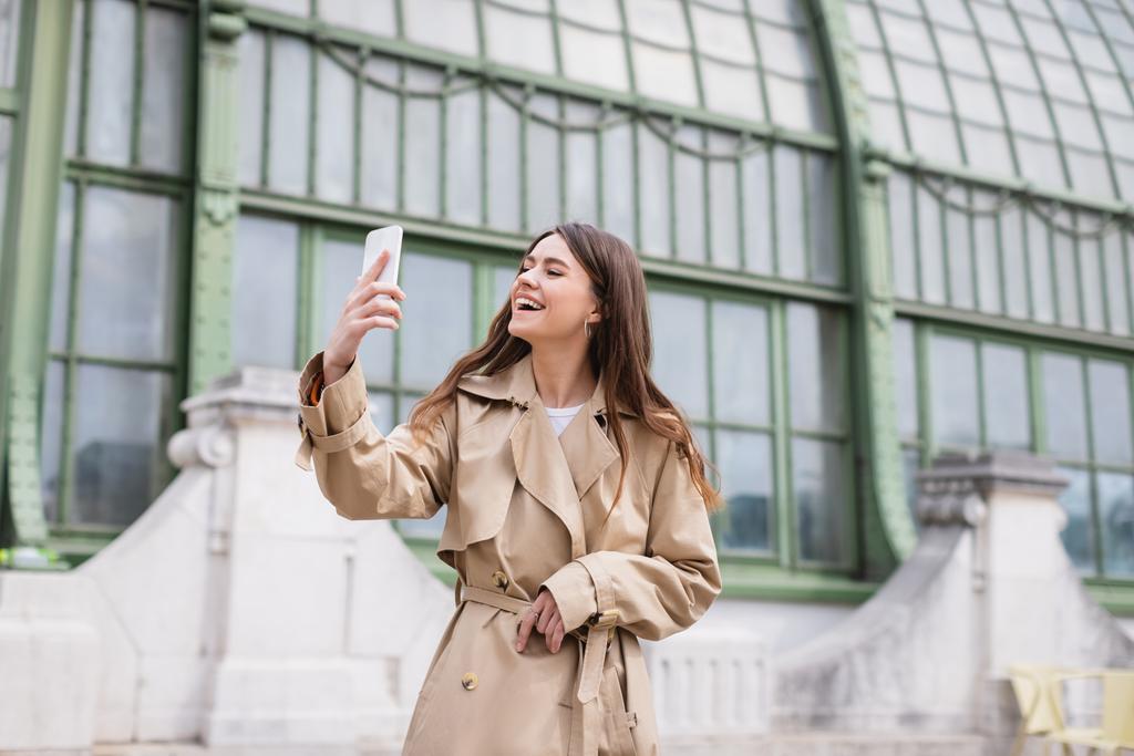 happy young woman in trench coat taking selfie near european building  - 写真・画像