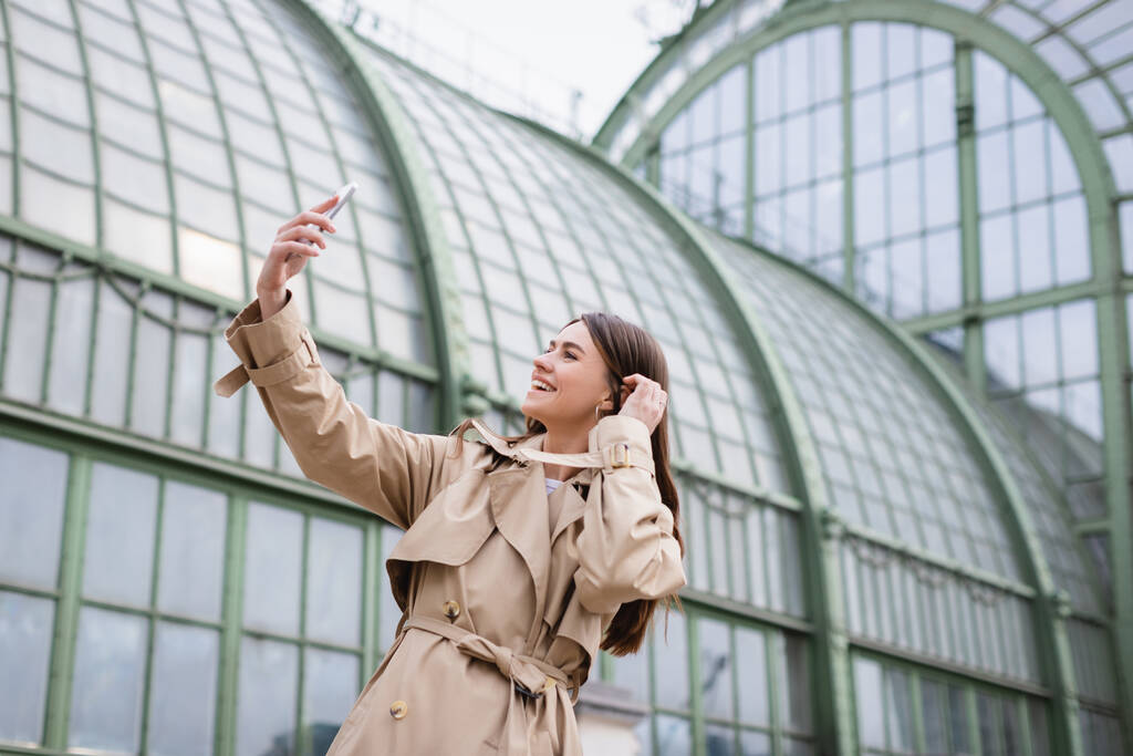 fröhliche junge Frau im Trenchcoat macht Selfie in der Nähe eines europäischen Gebäudes  - Foto, Bild