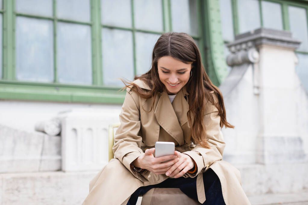 vrolijke vrouw in stijlvolle trench jas met behulp van mobiele telefoon in de buurt van Europese gebouw  - Foto, afbeelding