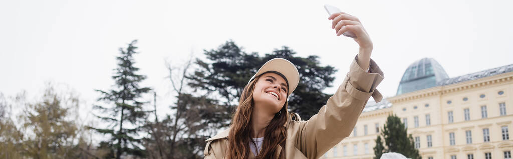 Ευτυχισμένη νεαρή γυναίκα με καμπαρντίνα και καπέλο του μπέιζμπολ λαμβάνοντας selfie στην ευρωπαϊκή πόλη, πανό - Φωτογραφία, εικόνα