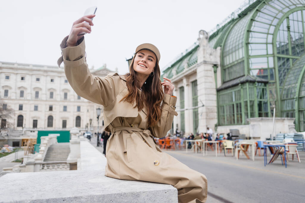 šťastná mladá žena ve stylové trenč kabát a baseballová čepice přičemž selfie  - Fotografie, Obrázek
