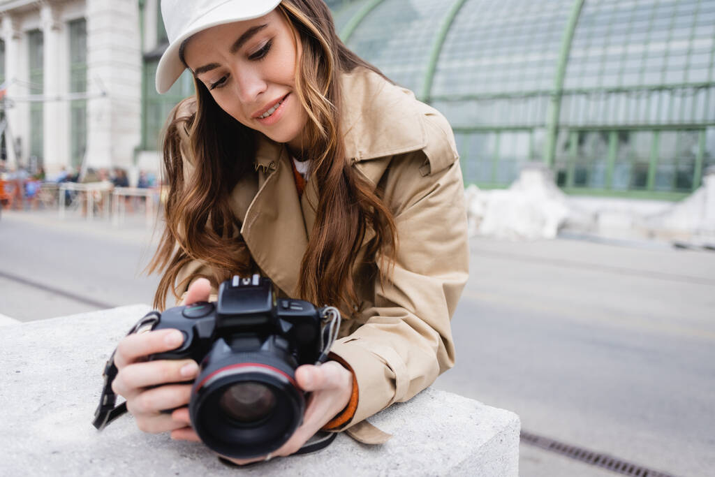 happy photographer in trench coat and baseball cap looking at digital camera  - Photo, Image