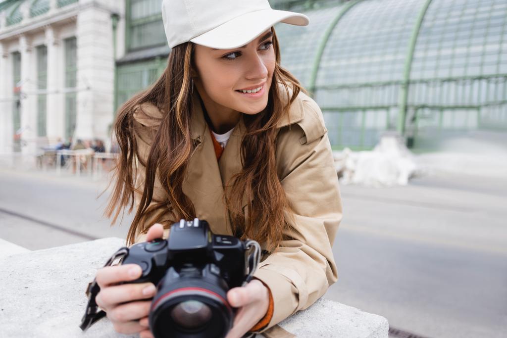 fotógrafo feliz en gabardina y gorra de béisbol sosteniendo cámara digital  - Foto, imagen