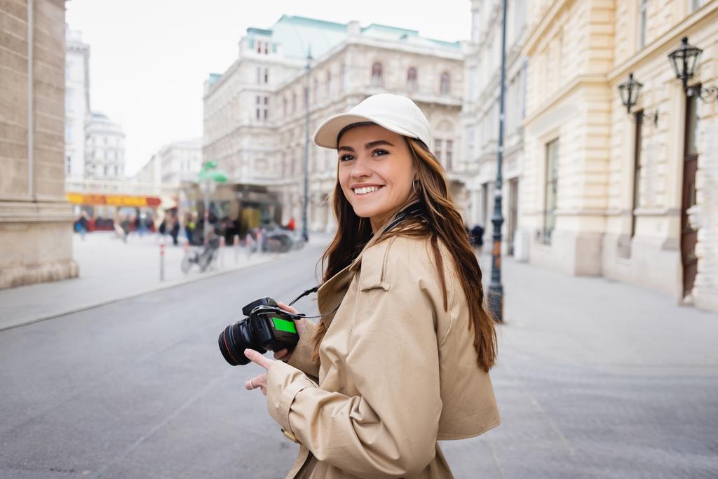 fröhliche Frau in Trenchcoat und Baseballkappe mit Digitalkamera auf der Straße einer europäischen Stadt - Foto, Bild