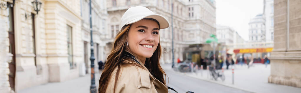 Trençkotlu ve beysbol şapkalı neşeli kadın Avrupa şehrinin caddesinde, afiş - Fotoğraf, Görsel