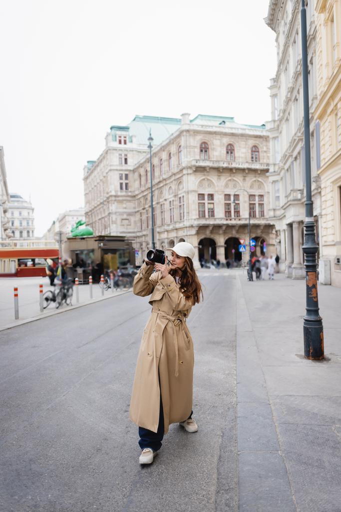 longitud completa de la mujer joven en abrigo de trinchera elegante y gorra de béisbol tomar fotos en la cámara digital en la ciudad europea - Foto, Imagen