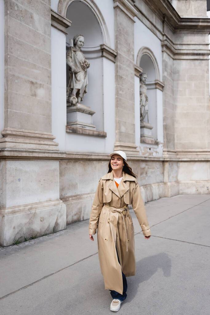 pełna długość szczęśliwej młodej kobiety w stylowym płaszczu i czapce baseballowej spacery po ulicy wiedeńskiej  - Zdjęcie, obraz
