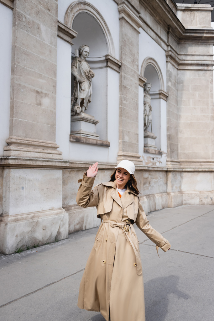 happy young woman in stylish trench coat and baseball cap walking and waving hand - 写真・画像