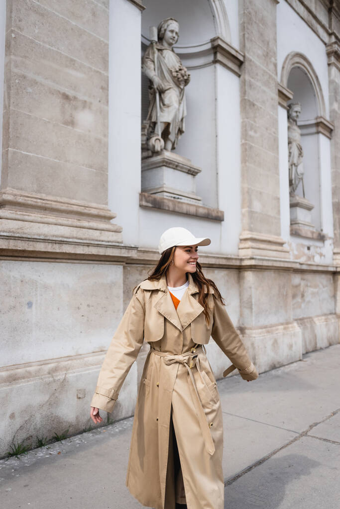 веселая молодая женщина в стильном плаще и бейсболке прогуливается по улице Вены  - Фото, изображение