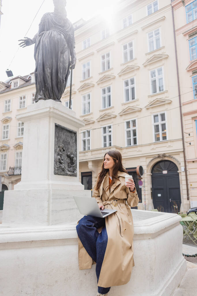 VIENA, ÁUSTRIA - MARÇO 30, 2022: mulher de casaco de trincheira sentada com laptop e segurando copo de papel na rua europeia  - Foto, Imagem