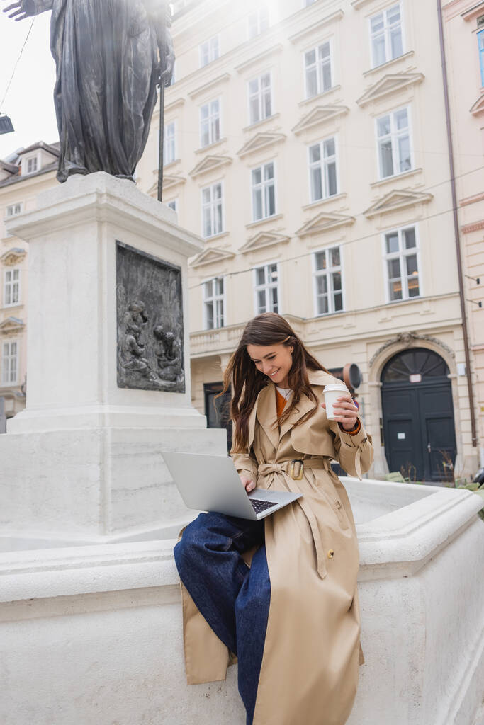happy young woman in stylish trench coat looking at laptop and holding paper cup on european street  - Photo, Image