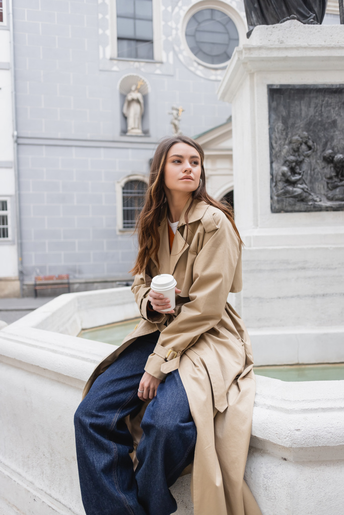 young woman in stylish trench coat holding paper cup on european street  - Photo, Image