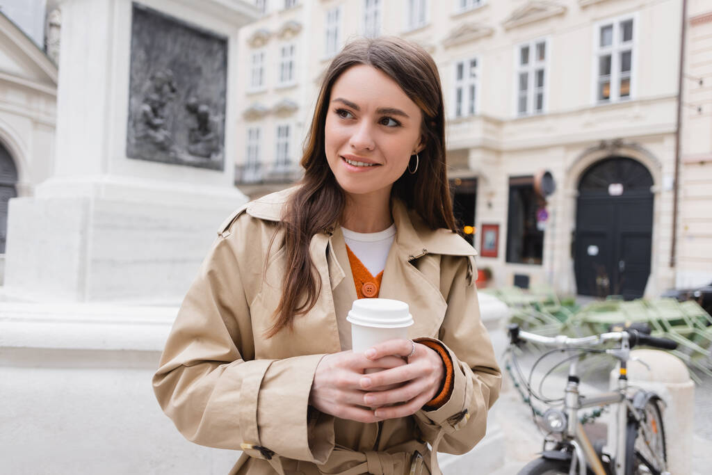 Улыбающаяся молодая женщина в стильном плаще держит бумажную чашку на европейской улице  - Фото, изображение