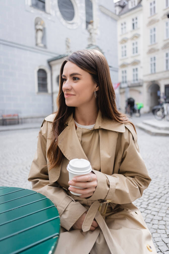 młoda uśmiechnięta kobieta w stylowym płaszczu, trzymająca papierowy kubek i siedząca na letnim tarasie w Europie - Zdjęcie, obraz
