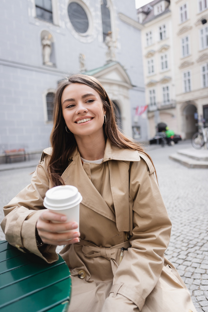 young cheerful woman in stylish trench coat holding paper cup on european street  - 写真・画像