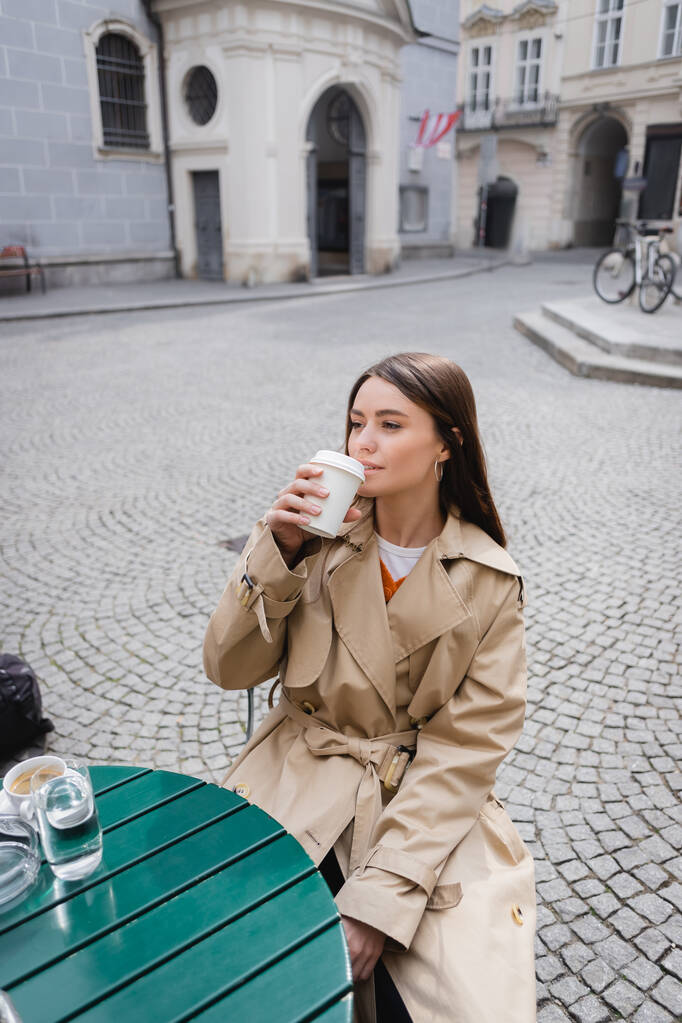 トレンチコートを着た若い女性がコーヒーを飲みながらカフェテラスに行き  - 写真・画像