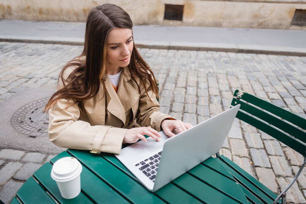 vista ad alto angolo di giovane donna in trench utilizzando il computer portatile vicino al caffè per andare  - Foto, immagini