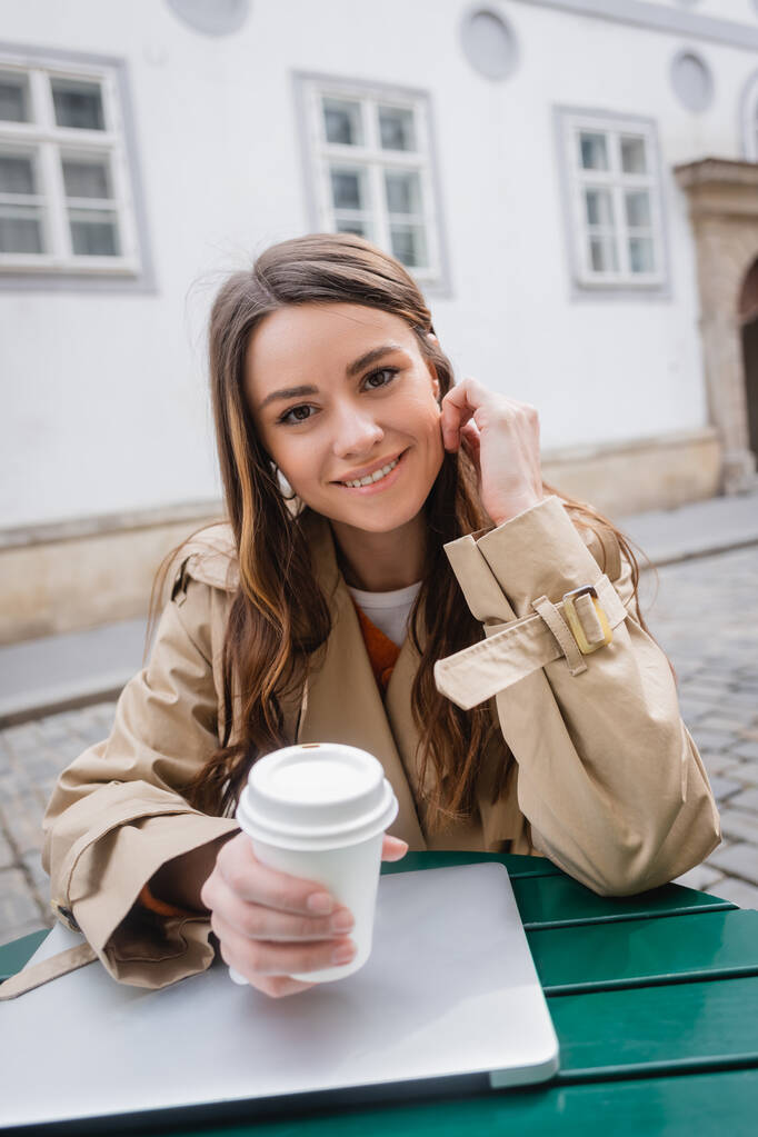 mujer joven feliz en gabardina de moda sosteniendo café para ir cerca de la computadora portátil en la terraza de verano  - Foto, imagen