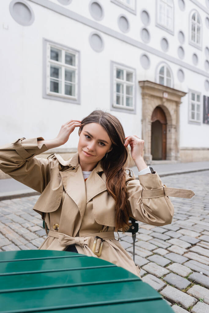 Nuori nainen trendikäs trendikäs trench takki säätämällä hiukset ja istuu kesäterassilla  - Valokuva, kuva