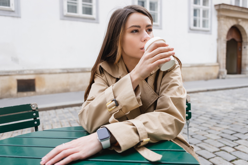mladá žena v módní trenč kabát drží papírový šálek a pití kávy jít na letní terasu  - Fotografie, Obrázek