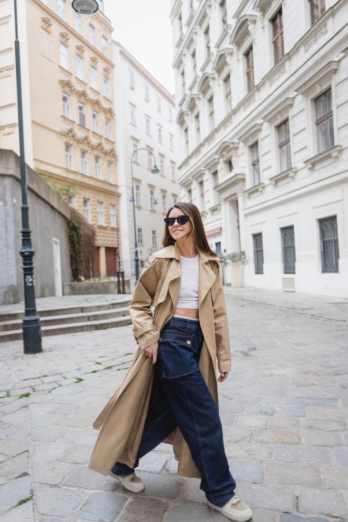 longitud completa de la joven alegre en gafas de sol de moda y abrigo de trinchera caminando en la calle de Viena - Foto, imagen