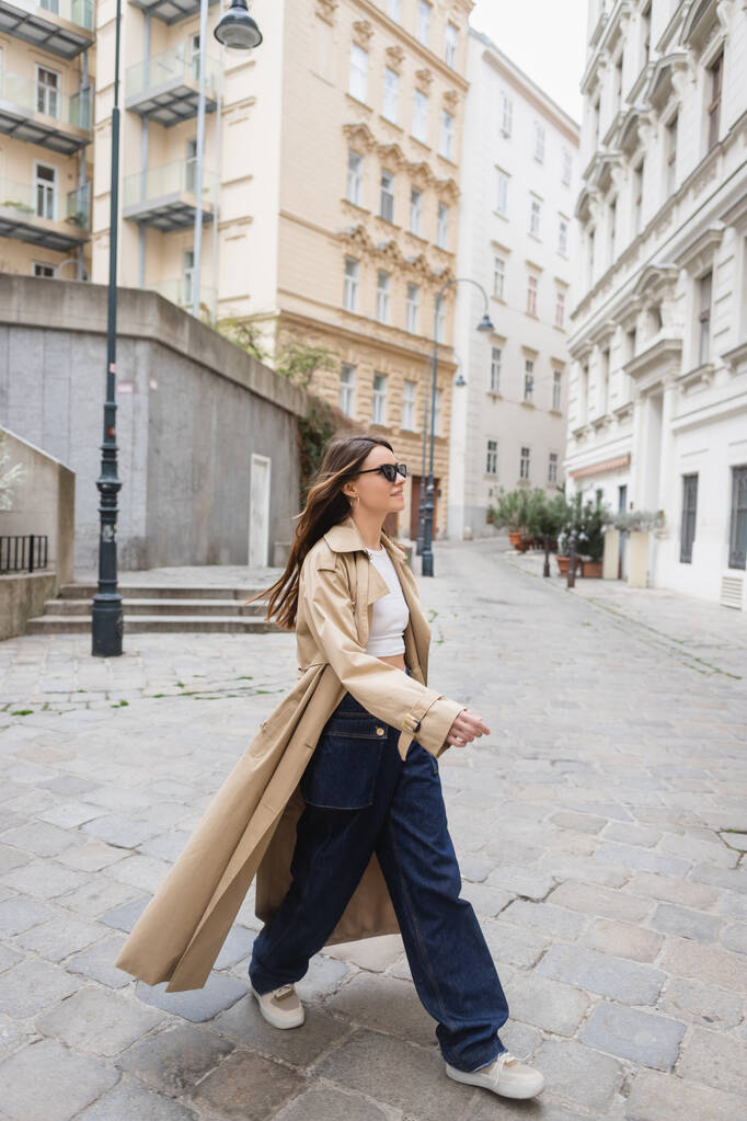 pleine longueur de jeune femme dans des lunettes de soleil à la mode et trench coat marche sur la rue de Vienne - Photo, image