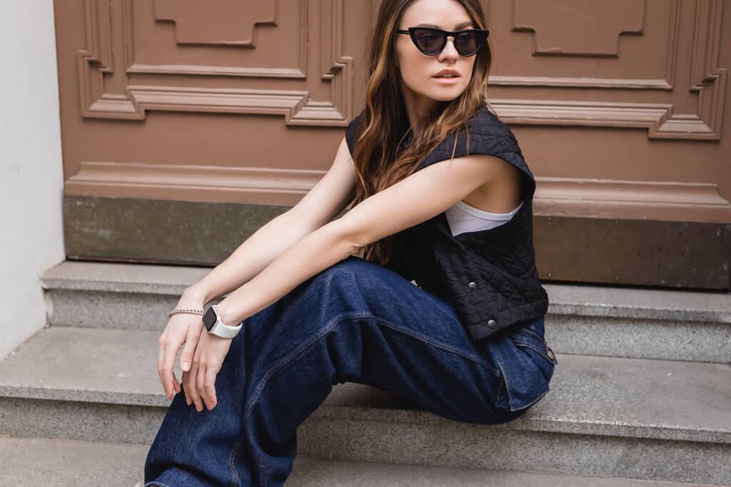 young woman in trendy sunglasses and sleeveless jacket sitting on stairs near entrance door  - Photo, Image