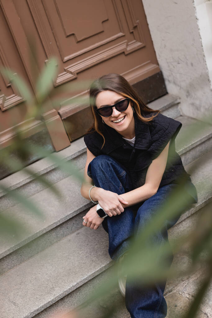 hoge hoek uitzicht van vrolijke jonge vrouw in trendy zonnebril en mouwloos jasje zittend op trap bij entree deur  - Foto, afbeelding