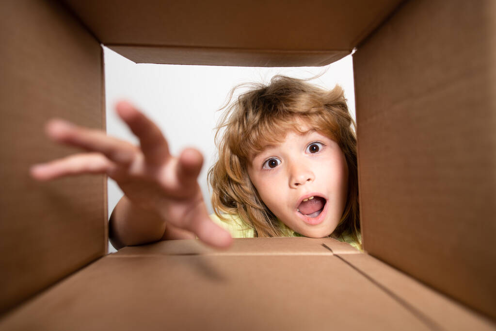 Dzieciak rozpakowuje i otwiera pudełko kartonowe, i patrzy do środka z zaskoczoną twarzą. Dziecko chłopiec patrząc zaskoczony w prezent. - Zdjęcie, obraz