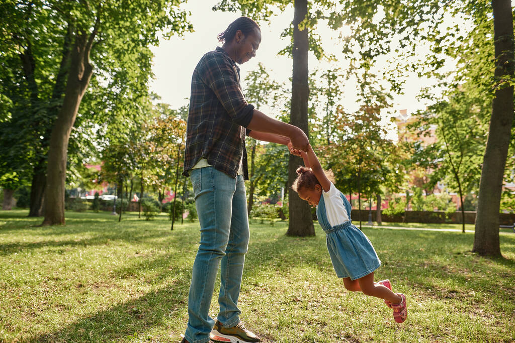 Black father spinning in hands daughter in park - Фото, изображение
