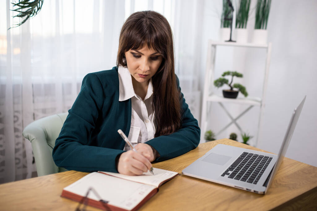 Portrait d'une fille dans une veste et chemise assise à une table et écrit des notes dans un carnet d'un ordinateur portable - Photo, image