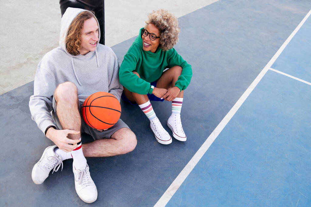 latin woman and caucasian man chatting happily sitting on the basketball court after the game, concept of friendship and urban sport in the street, copy space for text - Фото, изображение