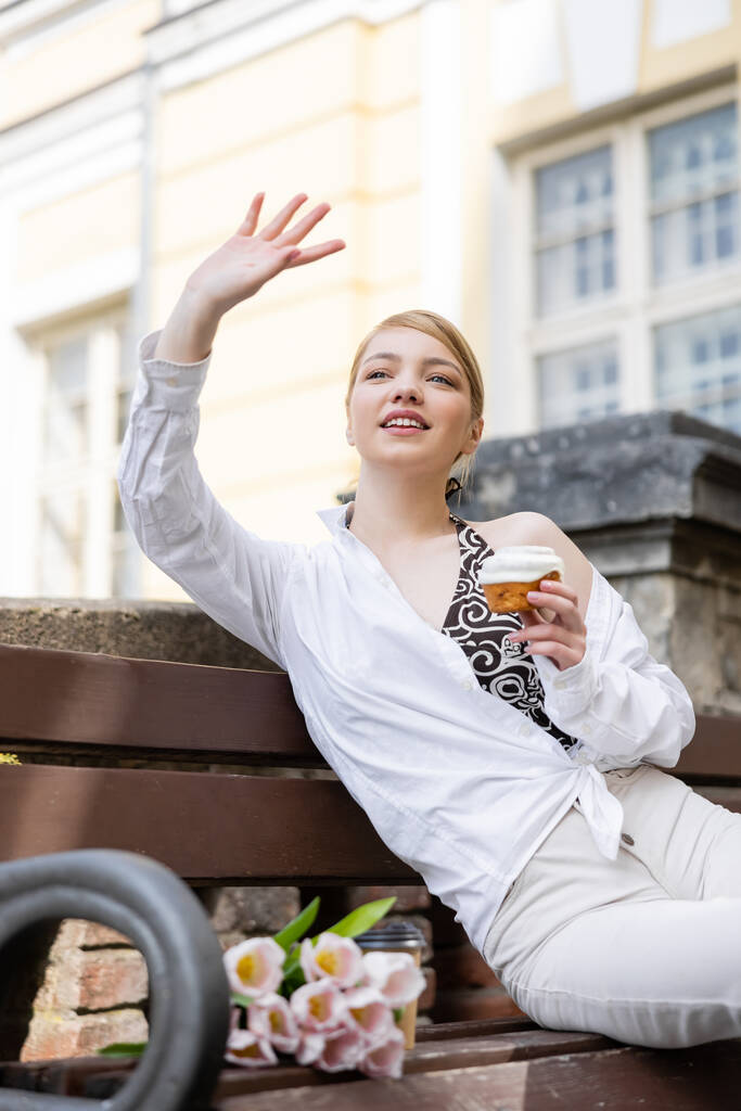 χαμογελαστή γυναίκα με cupcake κουνώντας το χέρι ενώ κάθεται στον πάγκο - Φωτογραφία, εικόνα