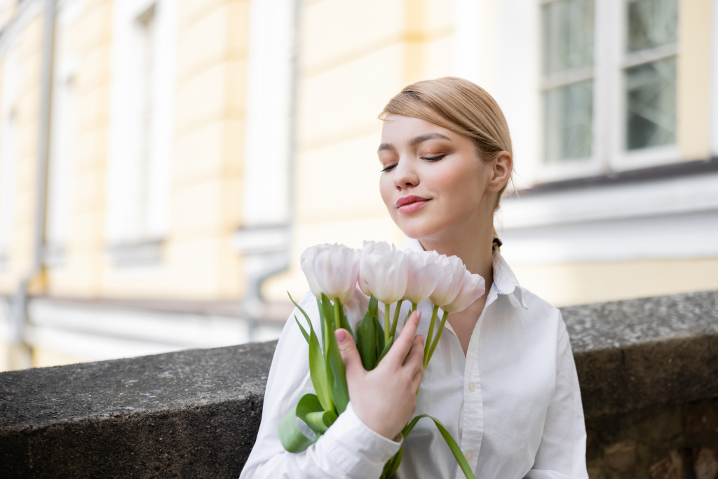 smiling blonde woman holding bouquet of white tulips on city street - Фото, изображение