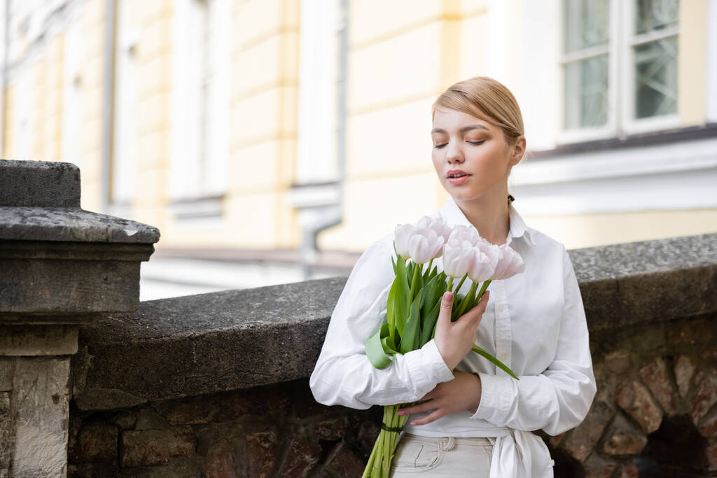 Frau in weißem Hemd steht mit Tulpenstrauß am Steinzaun - Foto, Bild