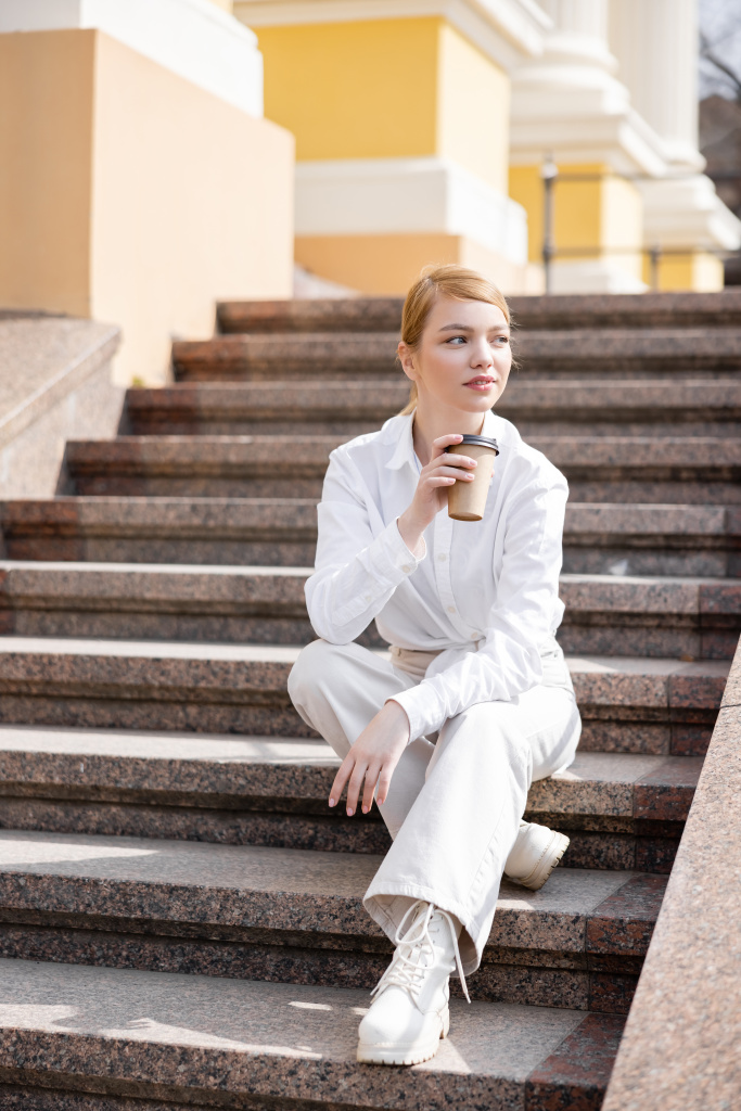 Ganzkörperansicht einer Frau in weißer Kleidung, die mit Kaffee to go auf einer Treppe sitzt - Foto, Bild