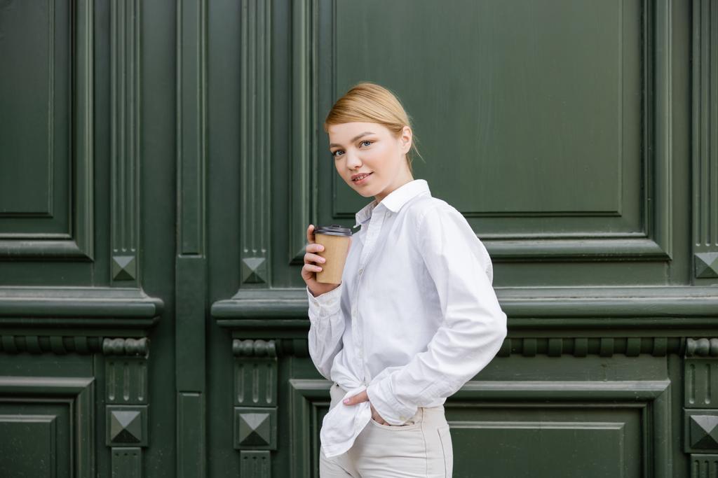 junge Frau im weißen Hemd steht mit der Hand in der Tasche und Kaffee in der Nähe der grauen Wand - Foto, Bild