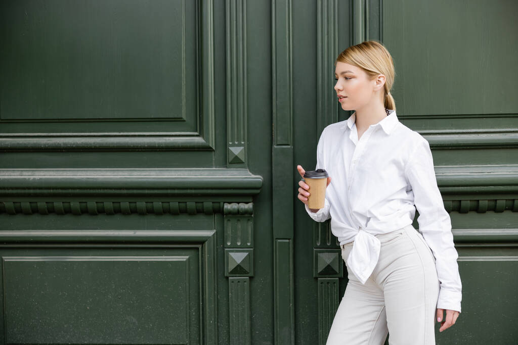 hezká žena v bílé košili a kalhotách stojící s papírovým kelímkem u šedé stěny - Fotografie, Obrázek