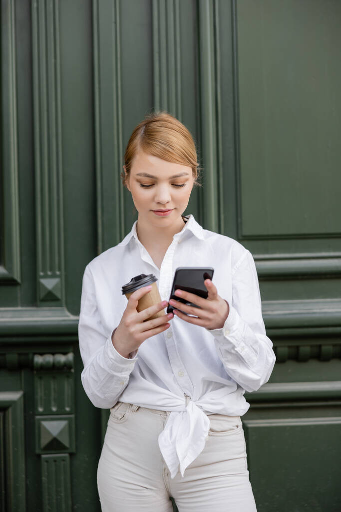 γυναίκα σε λευκό πουκάμισο κρατώντας χάρτινο κύπελλο και χρησιμοποιώντας κινητό τηλέφωνο κοντά σε γκρι τοίχο - Φωτογραφία, εικόνα