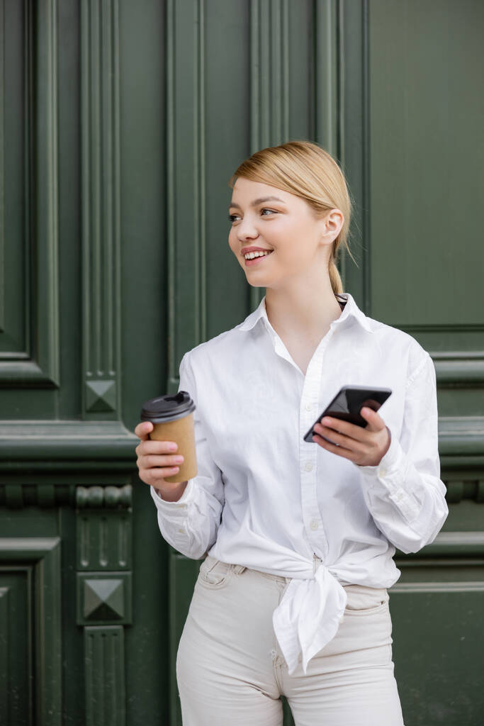 potěšená žena dívá pryč, zatímco stojí s chytrým telefonem a kávou jít do blízkosti šedé zdi - Fotografie, Obrázek