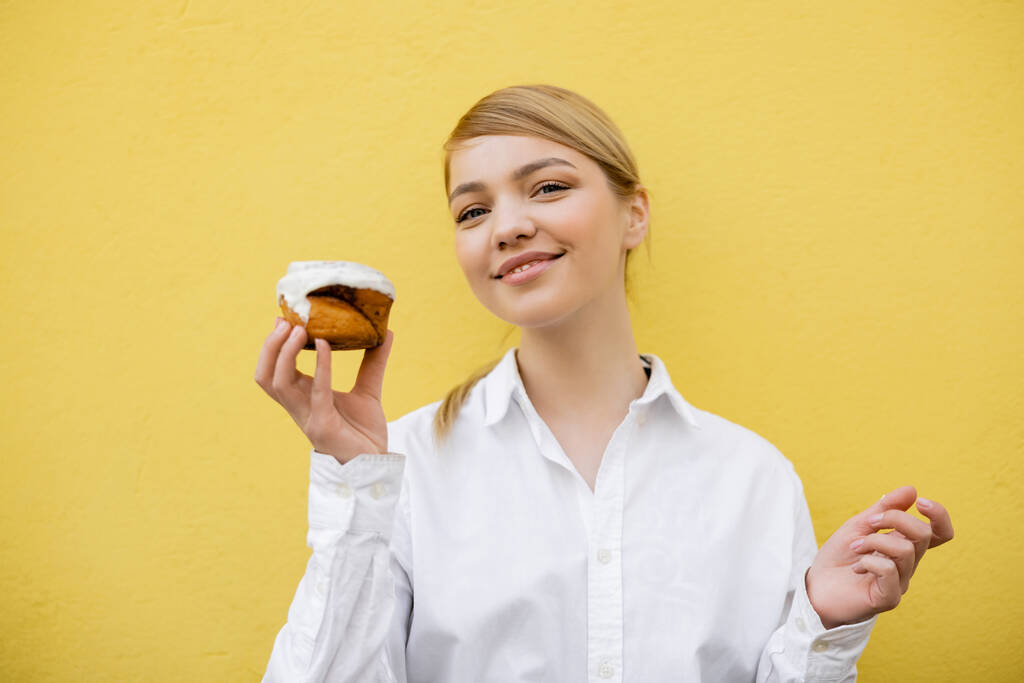 mujer feliz mirando a la cámara mientras sostiene cupcake con esmalte blanco aislado en amarillo - Foto, imagen