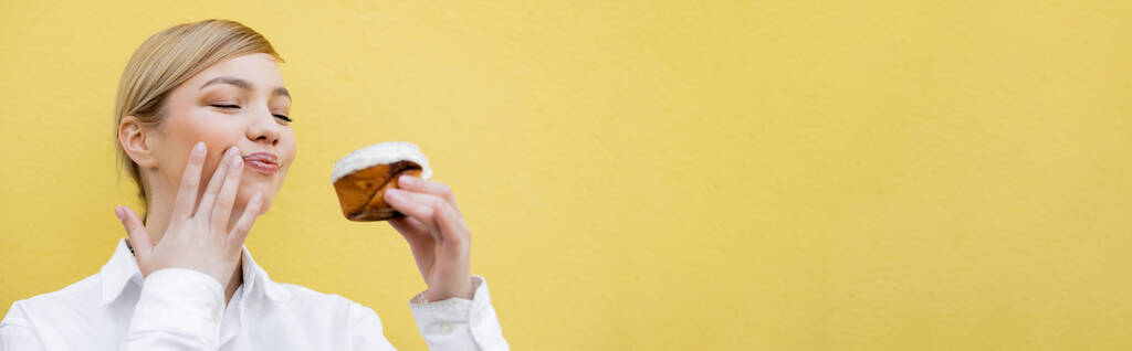 黄色のバナーで隔離されたおいしいカップケーキワイプ顔を持つ陽気な女性 - 写真・画像